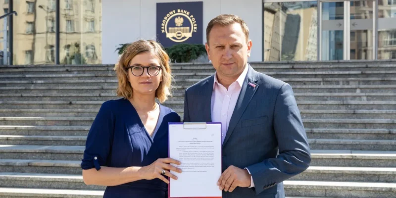 Na zdjęciu widoczni są dwaj ludzie pozujący na schodach przed budynkiem Narodowego Banku Polskiego, trzymający dokument.