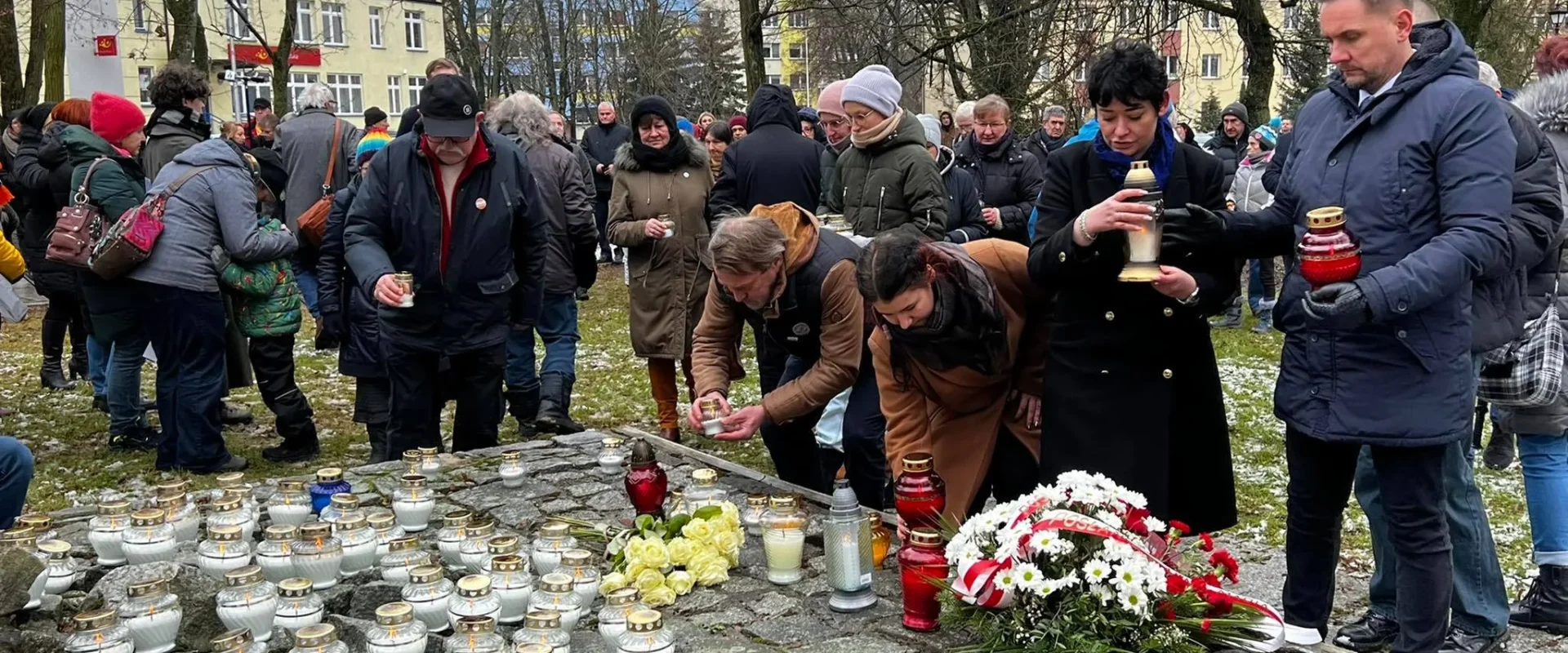 Zgromadzenie ludzi składających kwiaty i zapalających znicze w hołdzie w zamglonym parku.