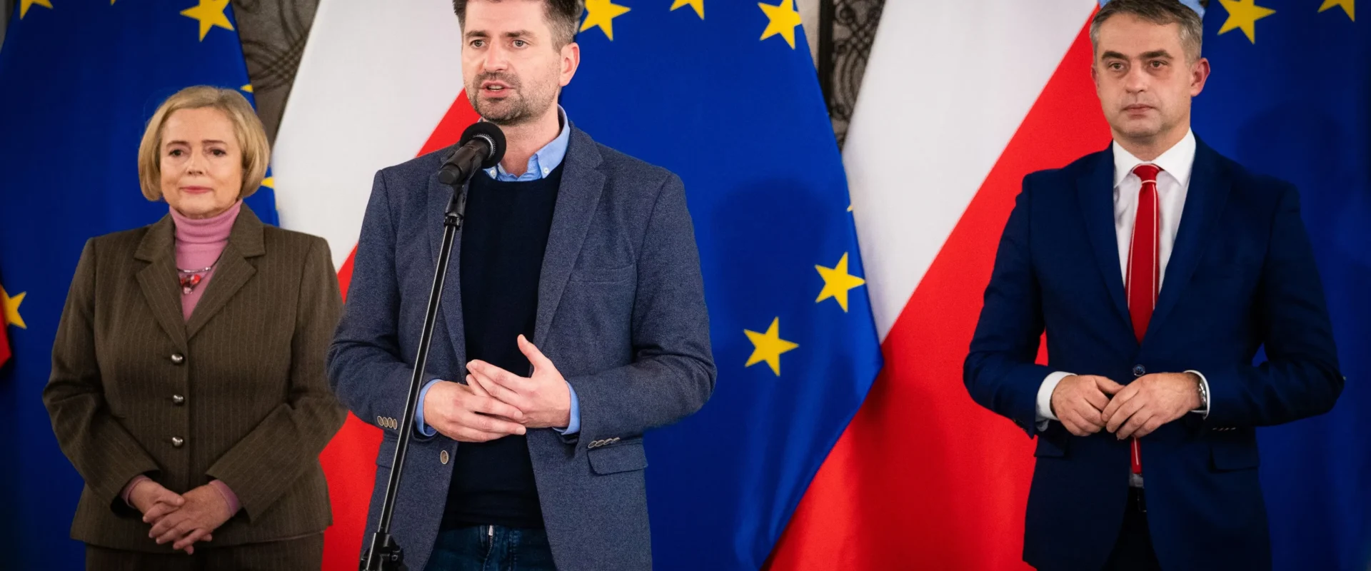 Trzy osoby stoją przed flagami Unii Europejskiej i Polski; środkowy mężczyzna przemawia do mikrofonu, po lewej stoi kobieta w brązowym garniturze, a po prawej mężczyzna w granatowym garniturze.