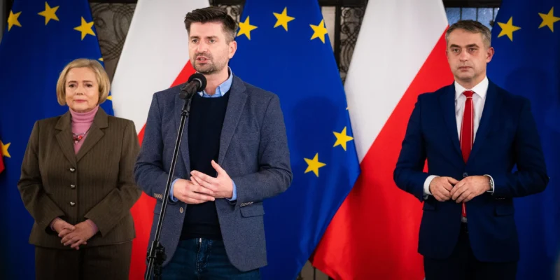 Trzy osoby stoją przed flagami Unii Europejskiej i Polski; środkowy mężczyzna przemawia do mikrofonu, po lewej stoi kobieta w brązowym garniturze, a po prawej mężczyzna w granatowym garniturze.