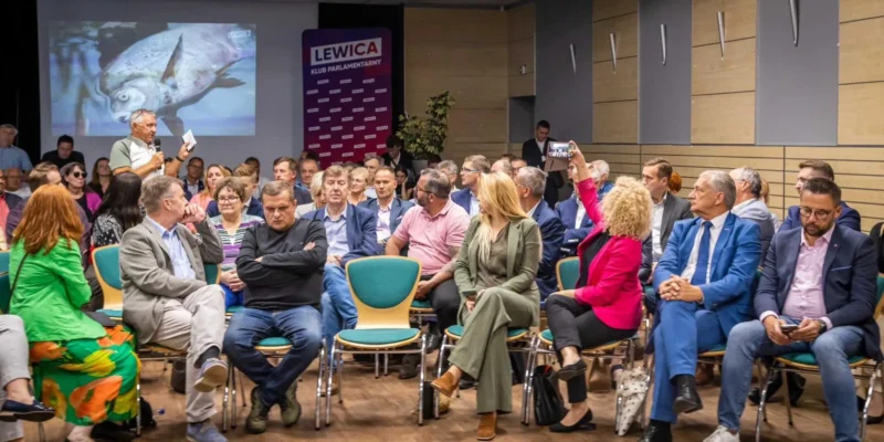 Na zdjęciu przedstawiono zebranie grupy ludzi siedzących w sali, z widoczną prezentacją na ścianie oraz jednym mężczyzną przemawiającym na pierwszym planie.