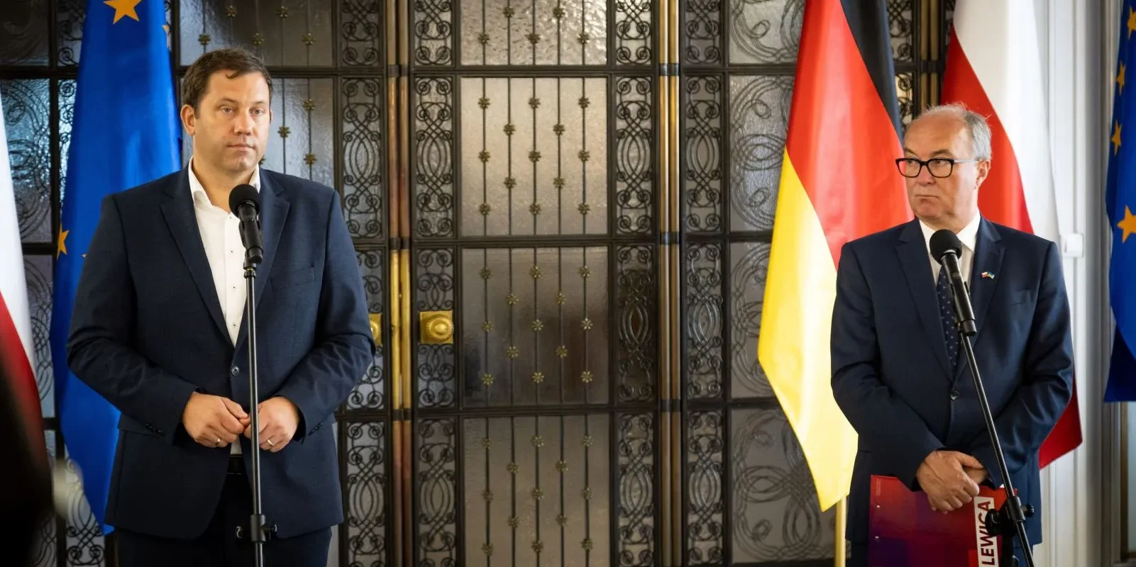 Na zdjęciu dwóch mężczyzn stoi przy mikrofonach w otoczeniu flagi niemieckiej, polskiej i unijnej, w czasie konferencji prasowej.