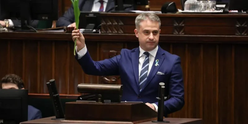 Mężczyzna w garniturze trzyma zieloną roślinę w podniesionej ręce, stojąc za biurkiem w sali obrad.