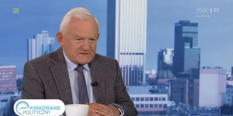 Mężczyzna w garniturze i krawacie siedzi za stołem w studiu telewizyjnym z filiżanką przed sobą, na ekranie widoczny napis "Kwadrans polityczny" i panorama miasta w tle.