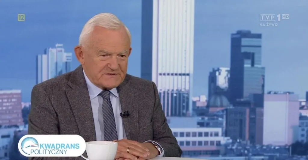 Mężczyzna w garniturze i krawacie siedzi za stołem w studiu telewizyjnym z filiżanką przed sobą, na ekranie widoczny napis "Kwadrans polityczny" i panorama miasta w tle.