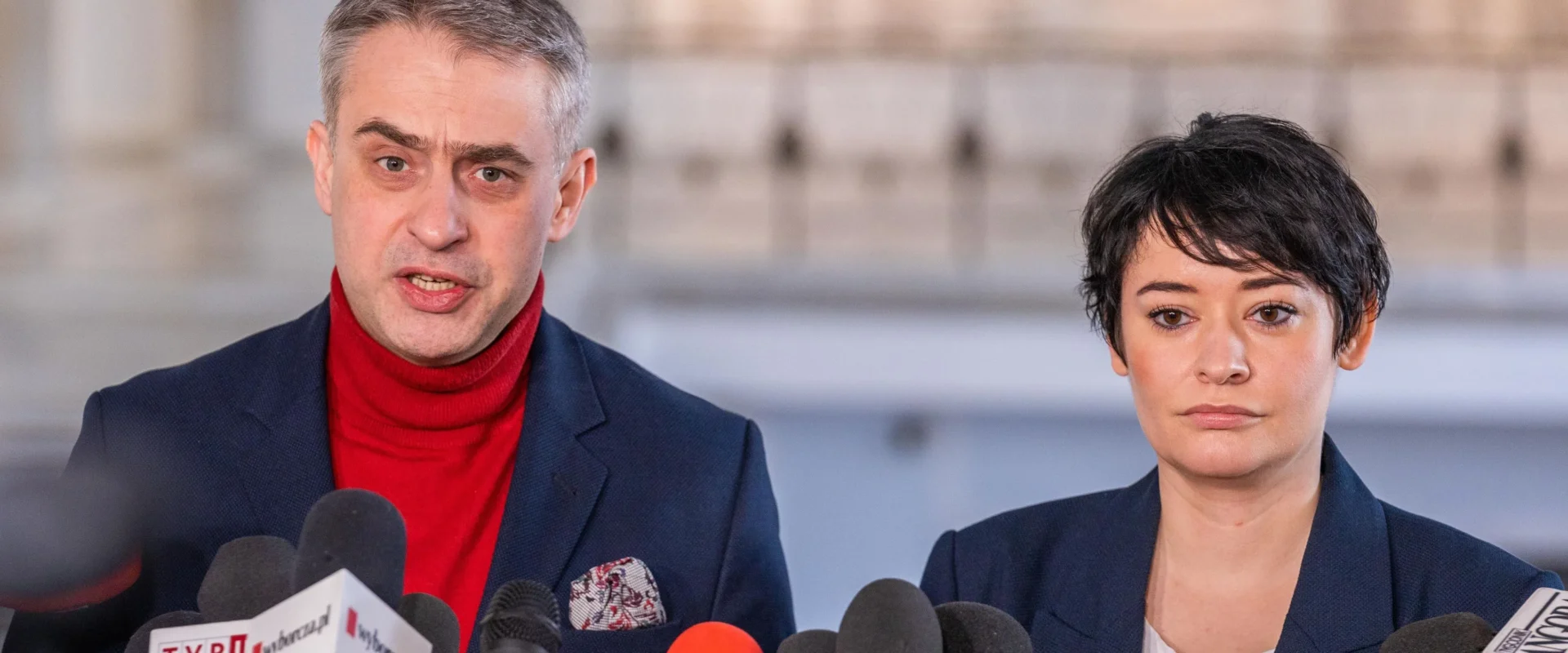 Dwóch polityków stoi za licznym szeregiem mikrofonów podczas konferencji prasowej; mężczyzna w czerwonym golfie i granatowej marynarce przemawia, obok niego stoi kobieta z krótkimi czarnymi włosami w granatowej marynarce.