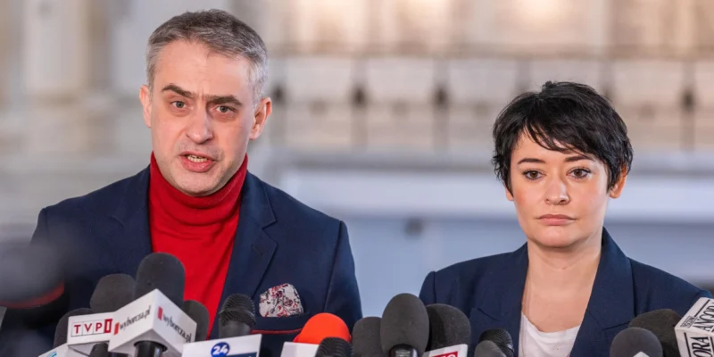 Dwóch polityków stoi za licznym szeregiem mikrofonów podczas konferencji prasowej; mężczyzna w czerwonym golfie i granatowej marynarce przemawia, obok niego stoi kobieta z krótkimi czarnymi włosami w granatowej marynarce.