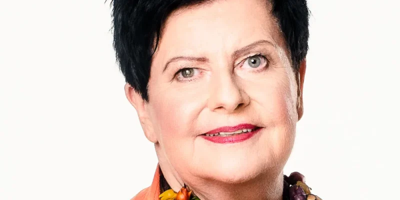 Portret kobiety o krótkich czarnych włosach, ubranej w pomarańczową bluzkę i wielobarwną, koralikową biżuterię na jasnym tle.