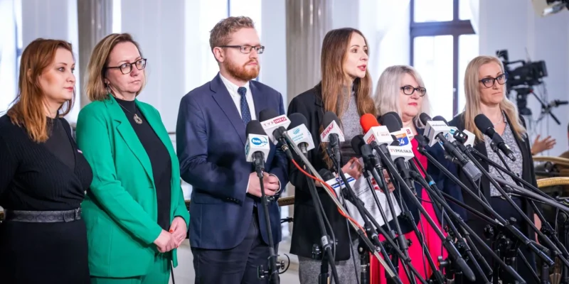 Na zdjęciu widać grupę kobiet i mężczyzn stojących przed mikrofonami podczas konferencji prasowej.