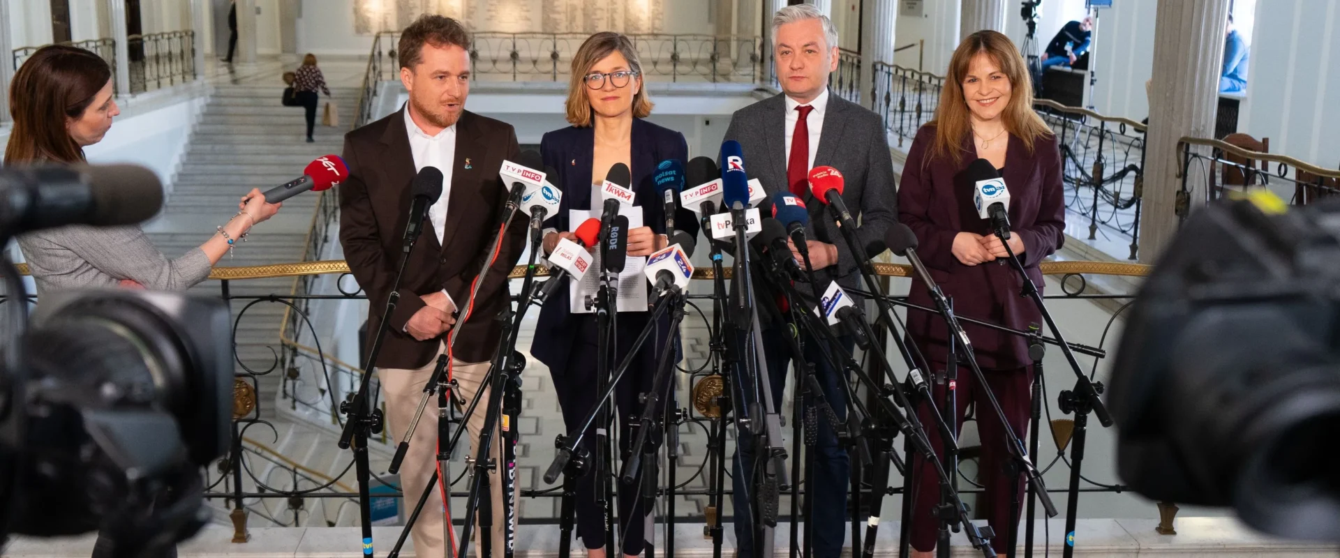 Grupa polityków lewicy podczas konferencji prasowej prezentuje ustawę antyflipperską, stojąc wśród licznych mikrofonów w holu budynku państwowego.