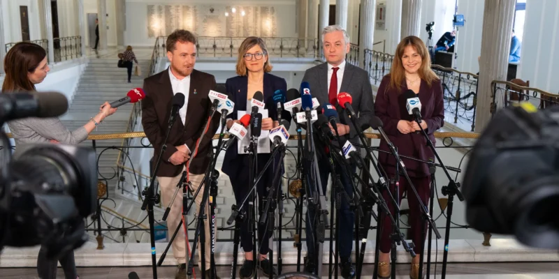 Grupa polityków lewicy podczas konferencji prasowej prezentuje ustawę antyflipperską, stojąc wśród licznych mikrofonów w holu budynku państwowego.