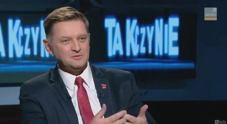 Mężczyzna w ciemnym garniturze i czerwonym krawacie gestykuluje podczas programu telewizyjnego, a za nim widać podświetlony ekran z napisem.