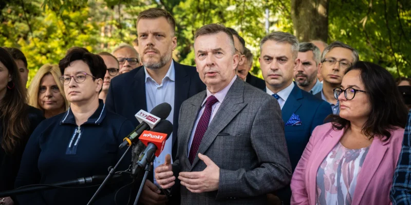 Grupa osób na konferencji prasowej na zewnątrz, w centrum mężczyzna w szarej kratkowanej marynarce przemawia do mikrofonów w otoczeniu zieleni.