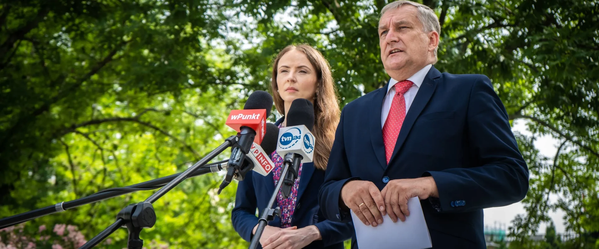 Na zewnątrz dwie osoby w garniturach udzielają konferencji prasowej, stojąc przy mikrofonach z logotypami mediów; w tle widać zielone drzewa.
