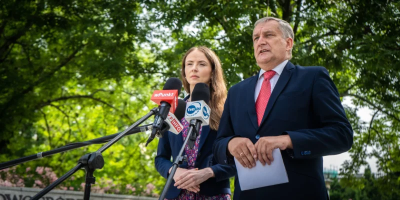 Na zewnątrz dwie osoby w garniturach udzielają konferencji prasowej, stojąc przy mikrofonach z logotypami mediów; w tle widać zielone drzewa.