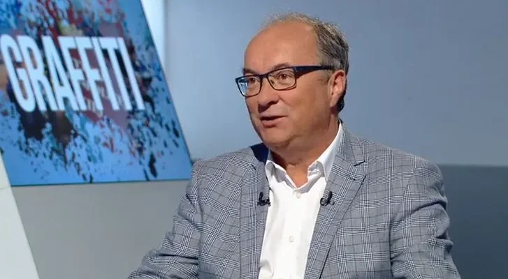 Mężczyzna w okularach, szarej kratkowanej marynarce i białej koszuli siedzi w studiu telewizyjnym, a w tle widać ekran z napisem GRAFFITI.