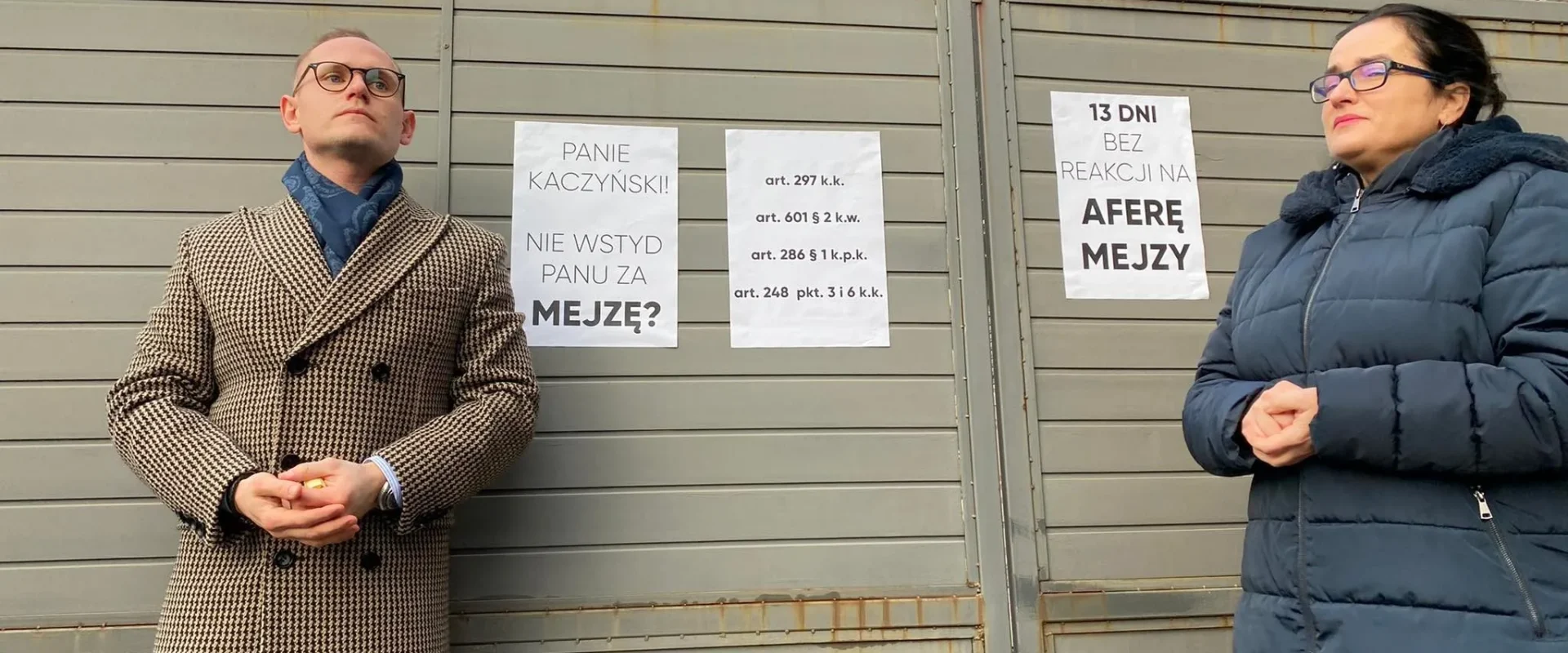 Dwóch uczestników konferencji Lewicy stoi przy metalowemu ogrodzeniu z plakatami dotyczącymi sprawy Łukasza Mejzy.