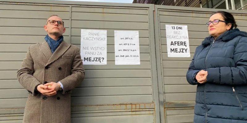 Dwóch uczestników konferencji Lewicy stoi przy metalowemu ogrodzeniu z plakatami dotyczącymi sprawy Łukasza Mejzy.
