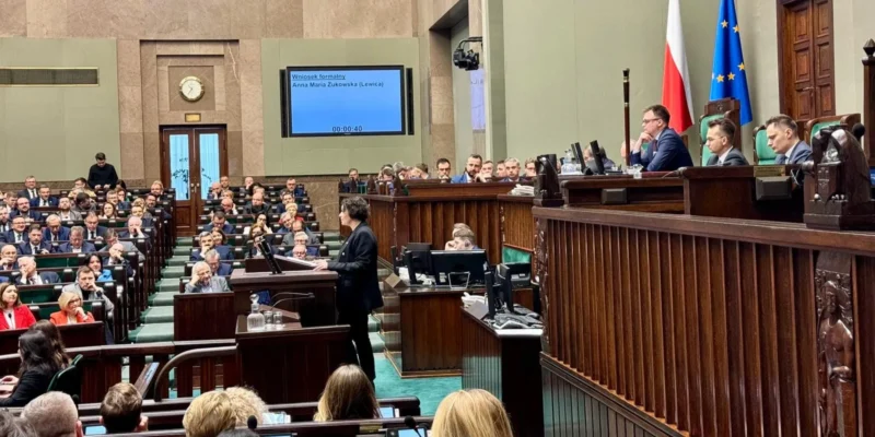 Osoba przemawiająca z mównicy w sali plenarnej Sejmu, otoczona zgromadzonymi posłami, z flagami Polski i Unii Europejskiej w tle i ekranem wyświetlającym komunikat o wniosku formalnym.