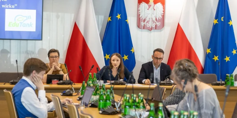 Na zdjęciu widoczna jest konferencja z udziałem kilku osób, w tym mówiącej kobiety, z flagami Polski i Unii Europejskiej w tle oraz zielonymi butelkami wody na stole.