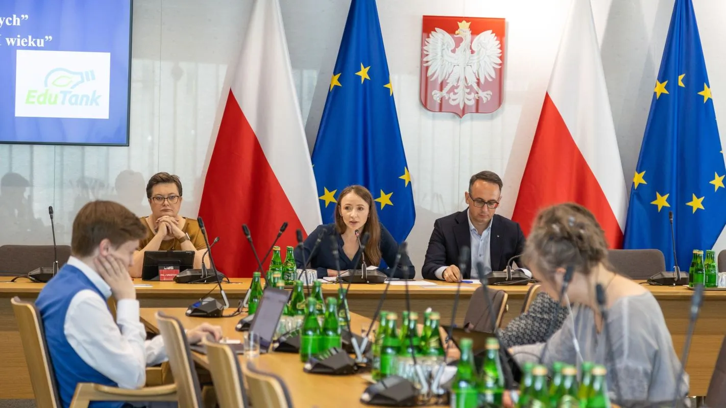 Na zdjęciu widoczna jest konferencja z udziałem kilku osób, w tym mówiącej kobiety, z flagami Polski i Unii Europejskiej w tle oraz zielonymi butelkami wody na stole.