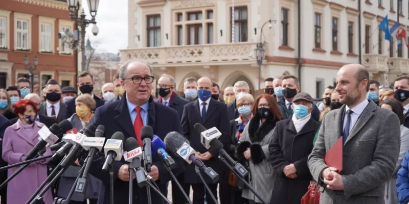 Polityk w granatowym garniturze i czerwonym krawacie przemawia do licznych mikrofonów podczas konferencji prasowej na placu, otoczony przez tłum zwolenników i zabytkowe budynki w tle.