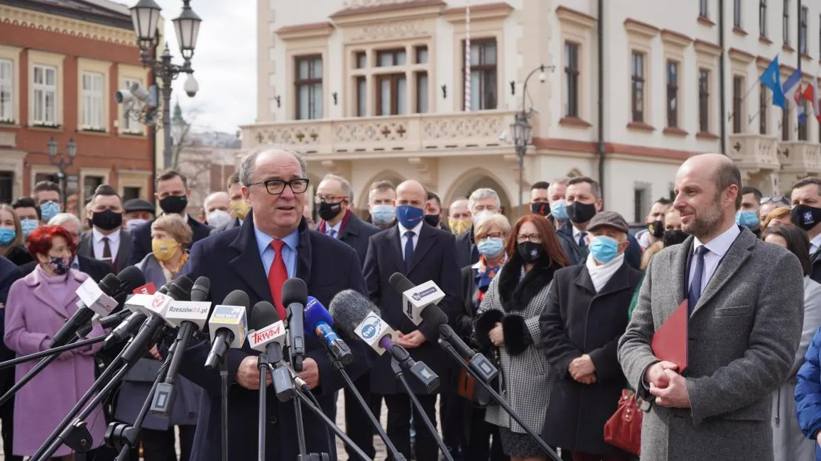 Polityk w granatowym garniturze i czerwonym krawacie przemawia do licznych mikrofonów podczas konferencji prasowej na placu, otoczony przez tłum zwolenników i zabytkowe budynki w tle.