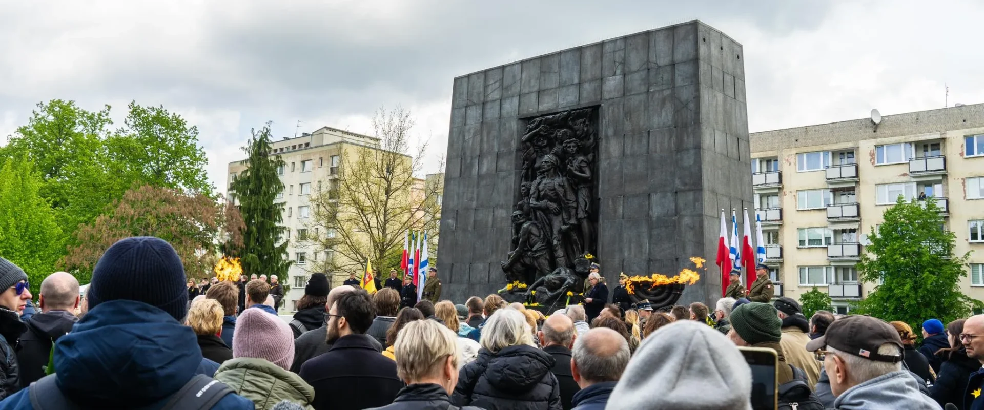 Tłum uczestników uroczystości 81. rocznicy powstania w getcie warszawskim przed monumentalnym pomnikiem z reliefem, obok płomieni ognia wiecznego i flag.