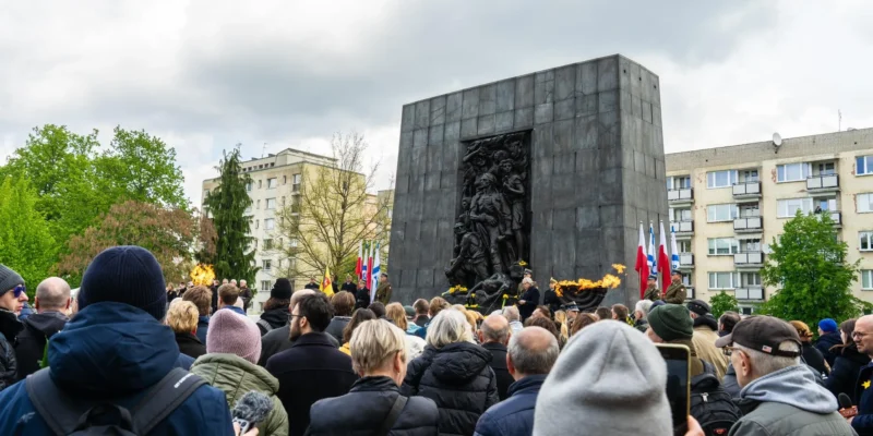 Tłum uczestników uroczystości 81. rocznicy powstania w getcie warszawskim przed monumentalnym pomnikiem z reliefem, obok płomieni ognia wiecznego i flag.