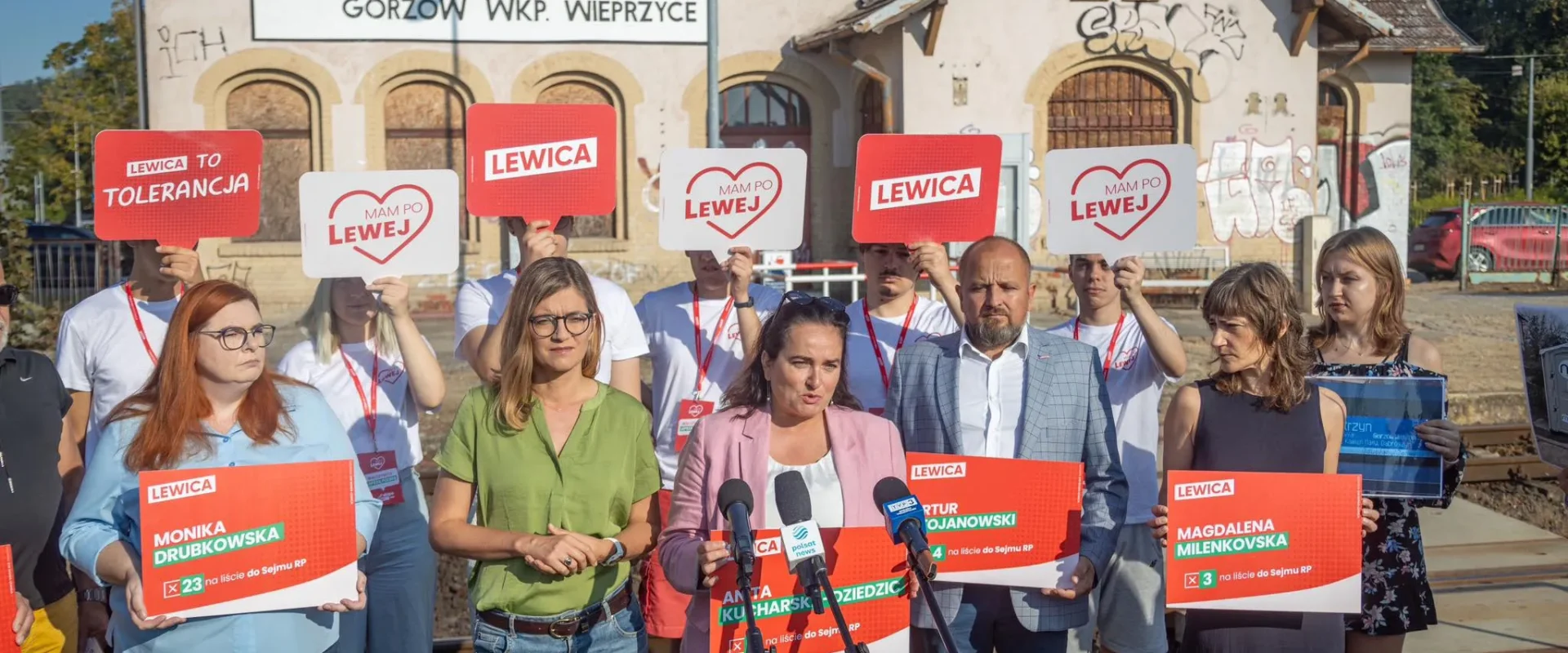 Grupa zwolenników Lewicy podczas konferencji prasowej przy torach, trzyma plakaty z napisem LEWICA i symbolem serca.