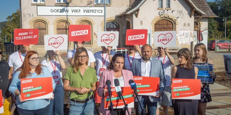Grupa zwolenników Lewicy podczas konferencji prasowej przy torach, trzyma plakaty z napisem LEWICA i symbolem serca.