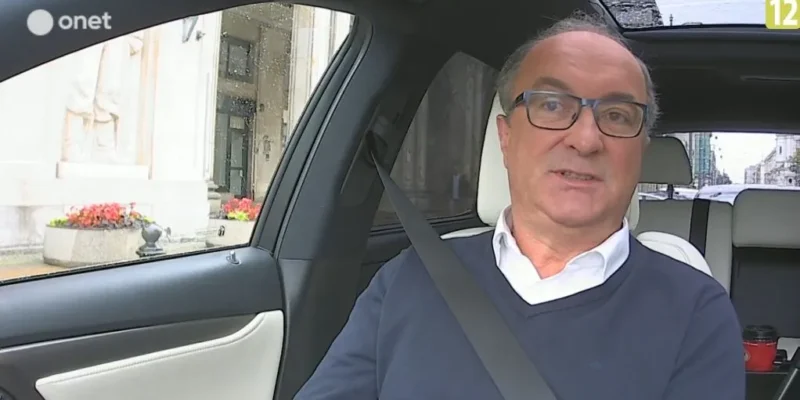 Mężczyzna w okularach siedzi przypięty pasem bezpieczeństwa w samochodzie i udziela wypowiedzi do kamery na temat obrony osób pracujących w okresie PRL (logo serwisu w lewym górnym rogu).