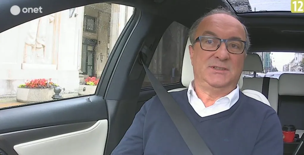 Mężczyzna w okularach siedzi przypięty pasem bezpieczeństwa w samochodzie i udziela wypowiedzi do kamery na temat obrony osób pracujących w okresie PRL (logo serwisu w lewym górnym rogu).