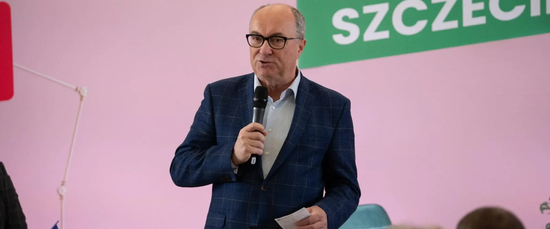 Mężczyzna w granatowym garniturze w kratę trzyma mikrofon i kartkę, przemawia na tle różowej ściany i zielonego baneru z napisem SZCZECIN.