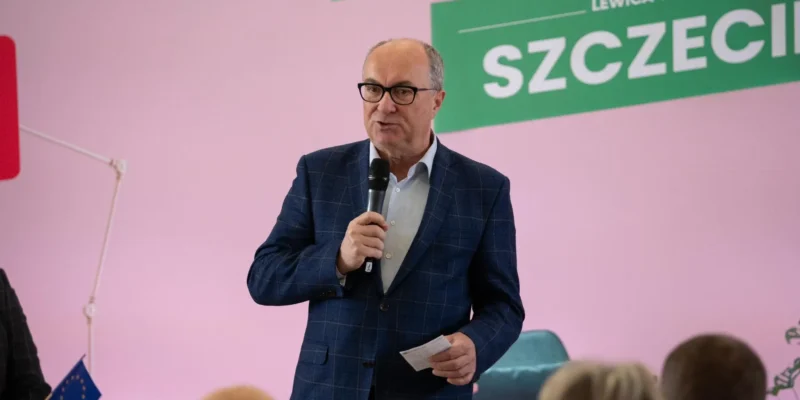 Mężczyzna w granatowym garniturze w kratę trzyma mikrofon i kartkę, przemawia na tle różowej ściany i zielonego baneru z napisem SZCZECIN.