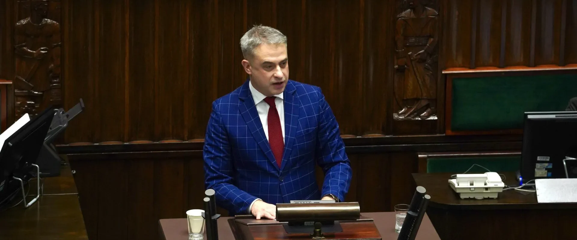 Mężczyzna w granatowym garniturze w kratkę i czerwonym krawacie stoi przy mównicy w sali obrad parlamentu.