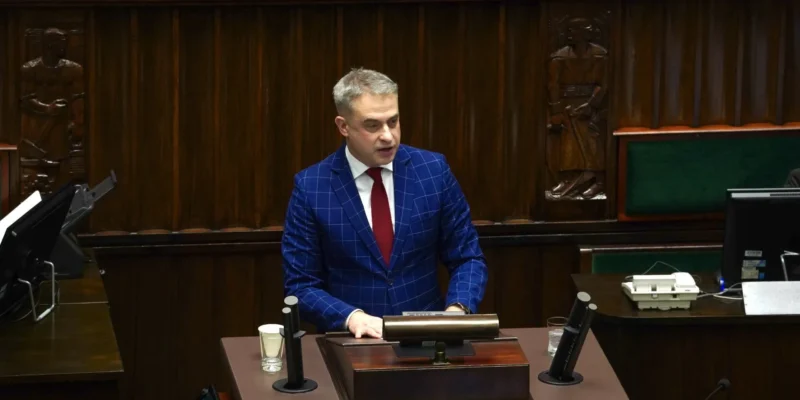 Mężczyzna w granatowym garniturze w kratkę i czerwonym krawacie stoi przy mównicy w sali obrad parlamentu.