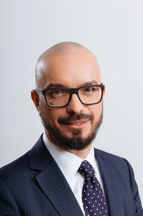 Portret studyjny łysiego mężczyzny z krótką brodą i okularami, w granatowym garniturze i krawacie w białe kropki, na jasnym tle.