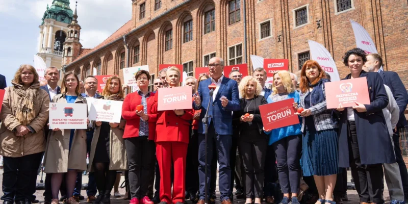 Grupa osób na placu przed historycznym budynkiem trzyma plakaty Lewicy z napisem Toruń, a na środku mężczyzna w błękitnym garniturze przemawia do mikrofonu.