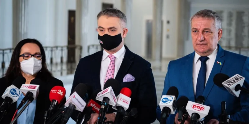 Na zdjęciu widać trójkę ludzi stojących przed mikrofonami podczas konferencji prasowej w eleganckim wnętrzu.