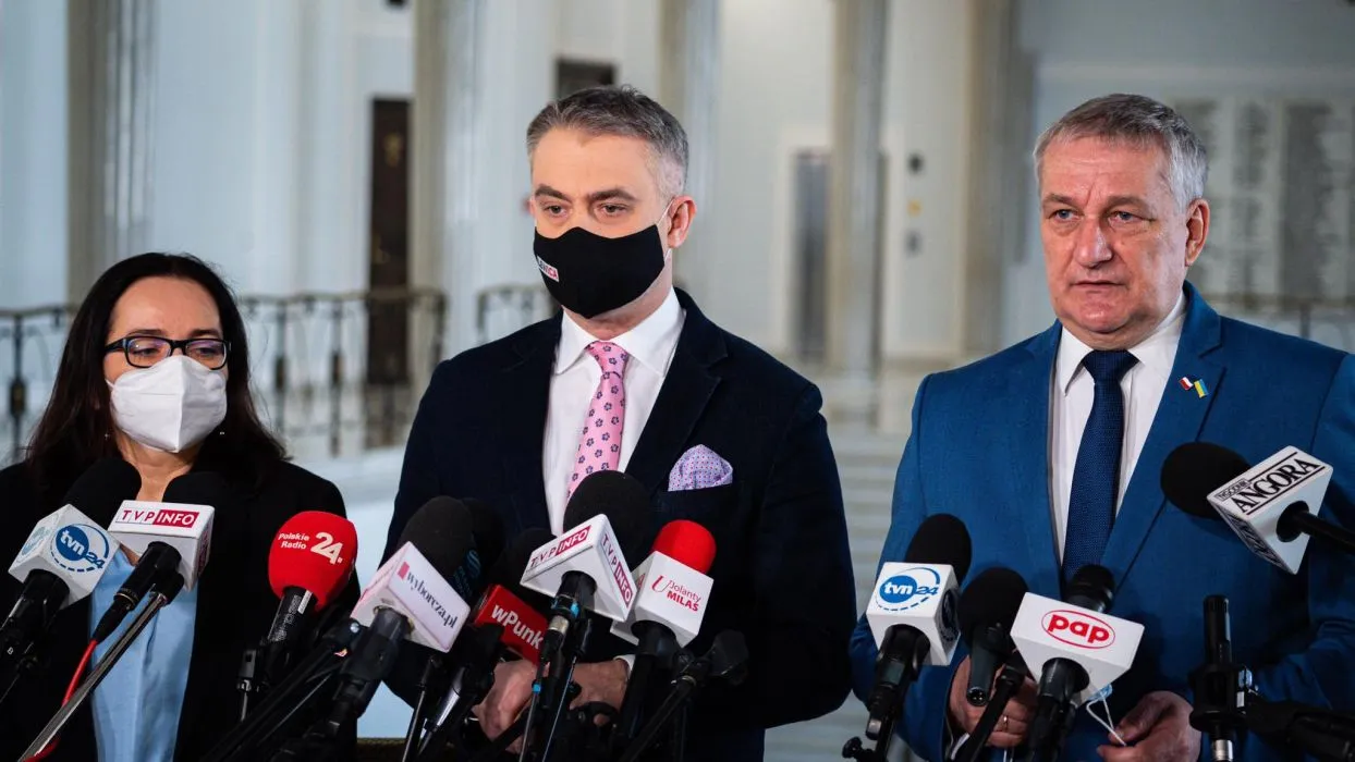 Na zdjęciu widać trójkę ludzi stojących przed mikrofonami podczas konferencji prasowej w eleganckim wnętrzu.