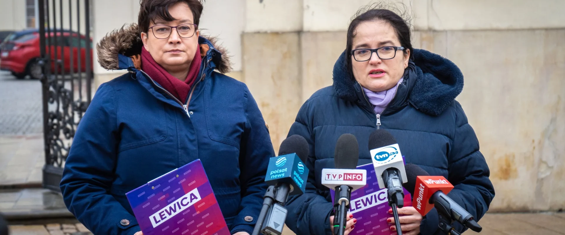 Dwie kobiety w zimowych kurtkach stoją przed zestawem mikrofonów mediów, trzymając fioletowe ulotki z napisem LEWICA podczas konferencji prasowej.