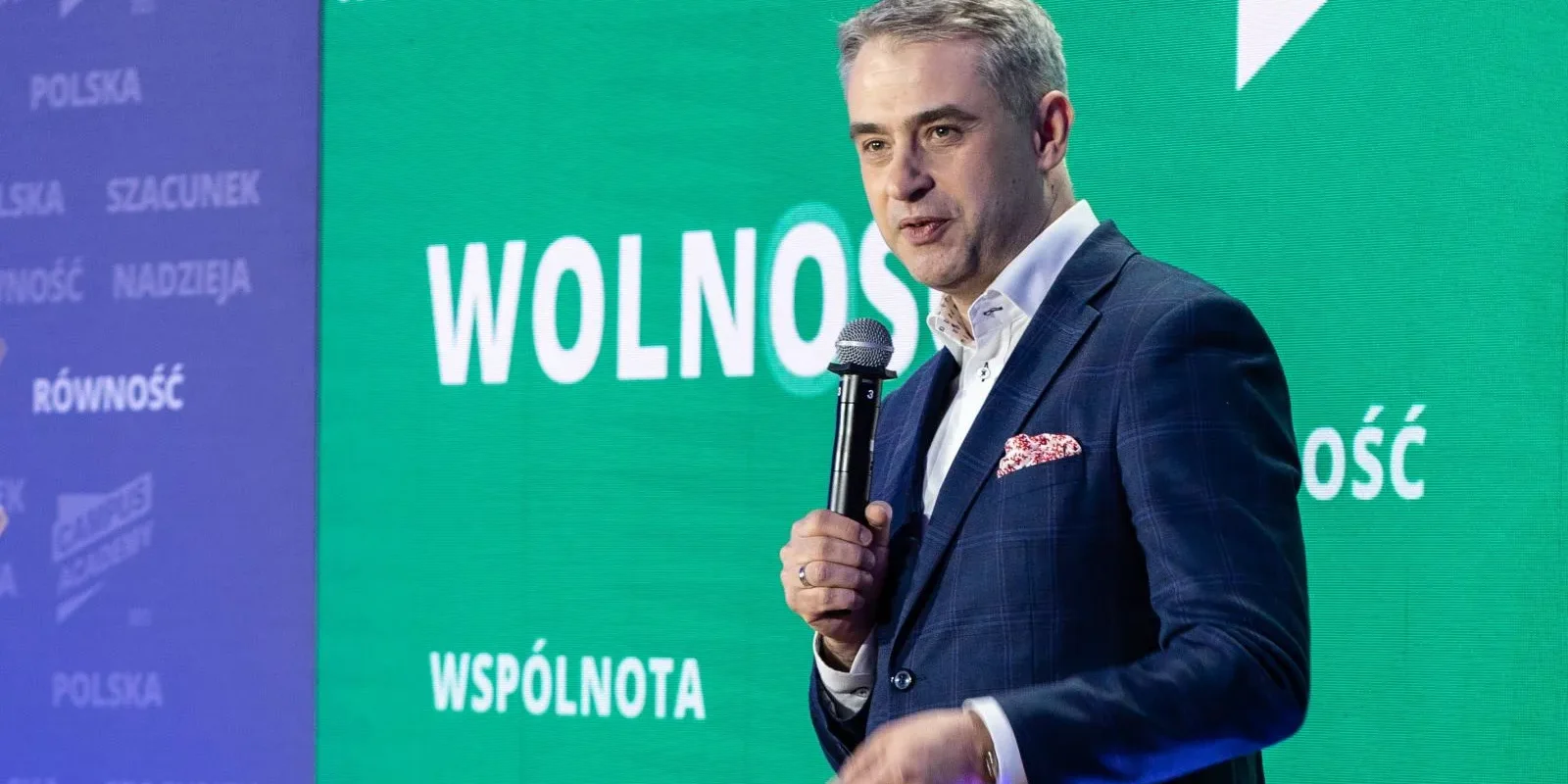 Mężczyzna w eleganckim garniturze przemawia do publiczności, trzymając mikrofon, w tle widoczny napis „WOLNOŚĆ” na zielonym tle.