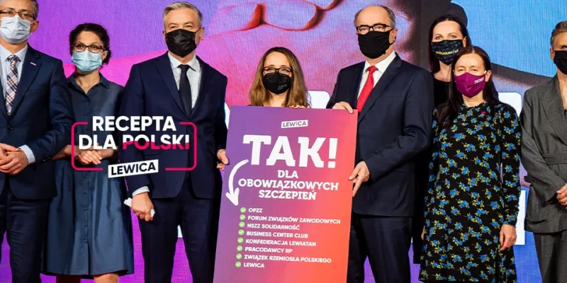 Grupa osób w garniturach i maseczkach na scenie trzyma plakat z napisem „TAK! DLA OBOWIĄZKOWYCH SZCZEPIEŃ” oraz baner z napisem „RECEPTA DLA POLSKI” i logiem Lewicy.