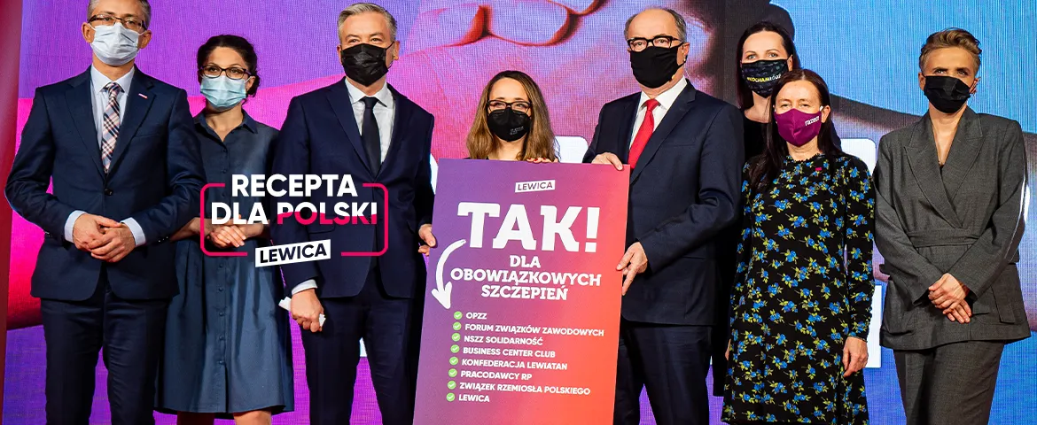 Grupa osób w garniturach i maseczkach na scenie trzyma plakat z napisem „TAK! DLA OBOWIĄZKOWYCH SZCZEPIEŃ” oraz baner z napisem „RECEPTA DLA POLSKI” i logiem Lewicy.