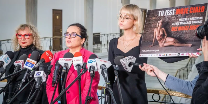 Trzy kobiety przemawiają na konferencji prasowej z okazji 16 dni przeciw przemocy wobec kobiet, a jedna z nich trzyma plakat z napisem „To jest moje ciało” i hasłem #TylkoTakOznaczaZgode.