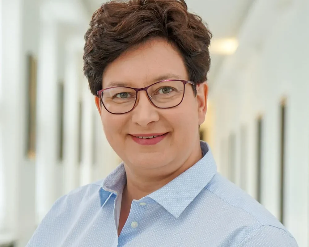 Portret uśmiechniętej kobiety w niebieskiej koszuli, stojącej w jasnym korytarzu.