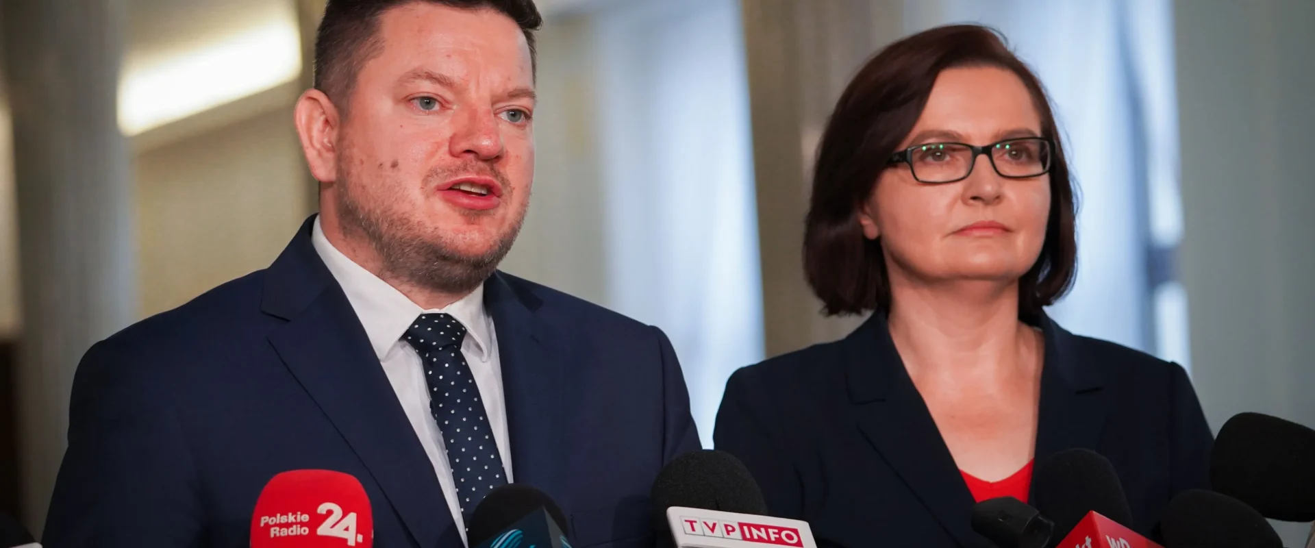 Na zdjęciu widoczna jest para mówiących do mikrofonów osób w formalnych ubraniach, stojących przed prasą.