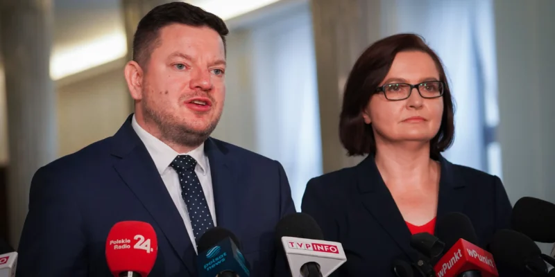 Na zdjęciu widoczna jest para mówiących do mikrofonów osób w formalnych ubraniach, stojących przed prasą.