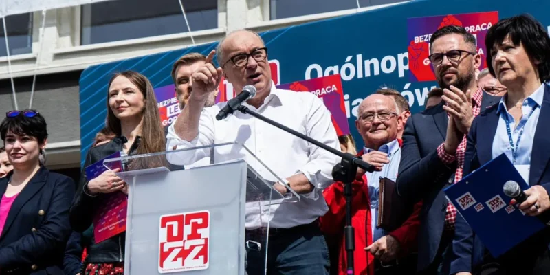 Polityk przemawia z mikrofonem na podium podczas wiecu, otoczony innymi uczestnikami i plakatami w tle.